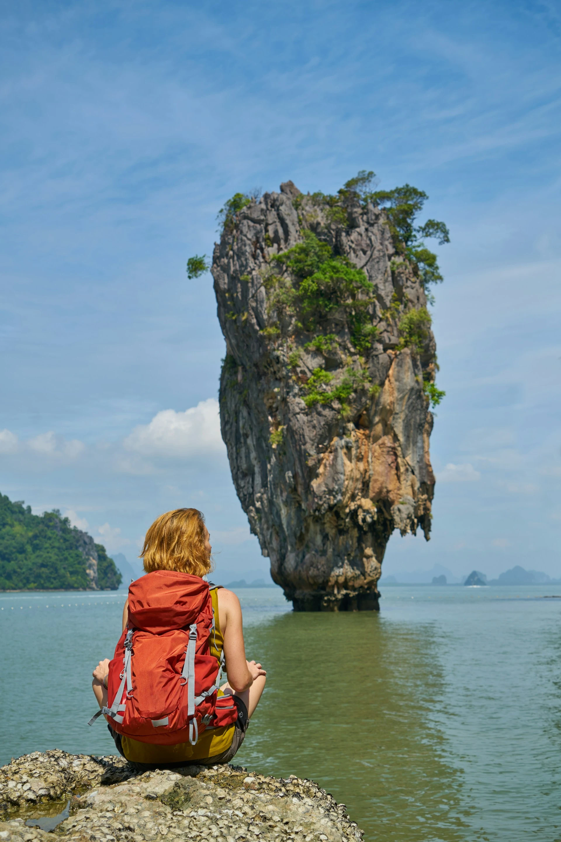 Traveller with a red backpack sitting on a rock facing the Ko Tapu limestone pinnacle rising from the sea at James Bond Island
