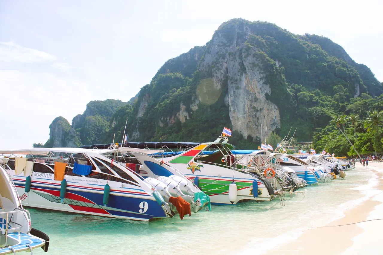 Row of colourful speedboats moored in shallow turquoise water along a beach below a jungle-covered limestone cliff in the Phi Phi Islands