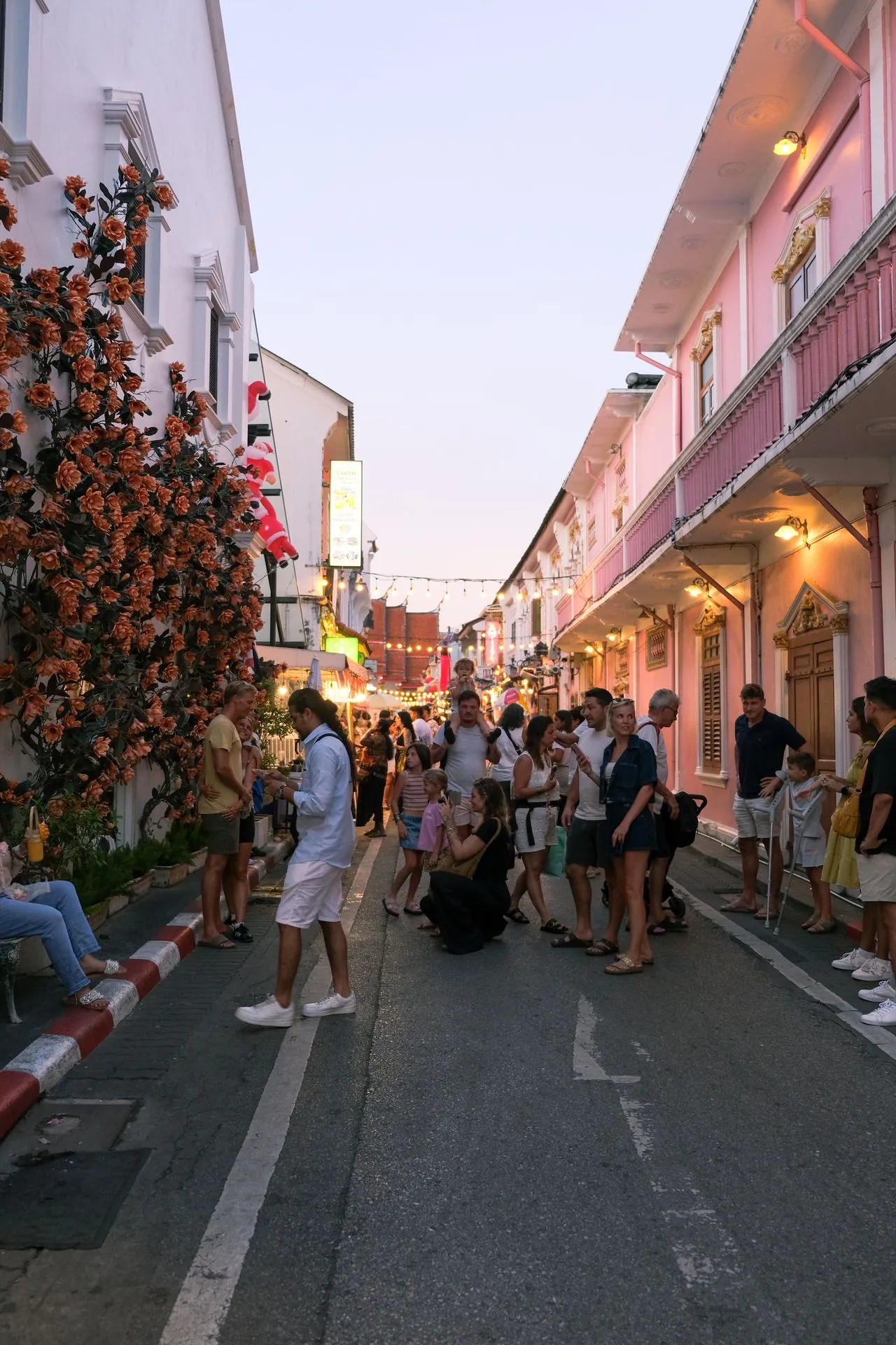 Crowds on a Phuket Old Town street at dusk between pastel pink and white Sino-Portuguese shophouses hung with string lights
